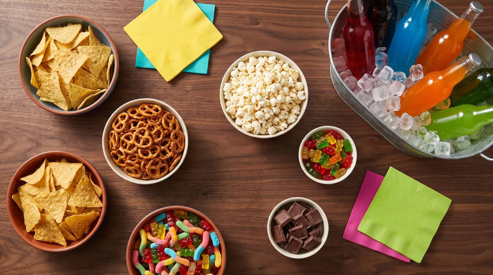 Overhead view of a party snack table with chips, pretzels, popcorn, candy and a tub of iced sodas on dark wood