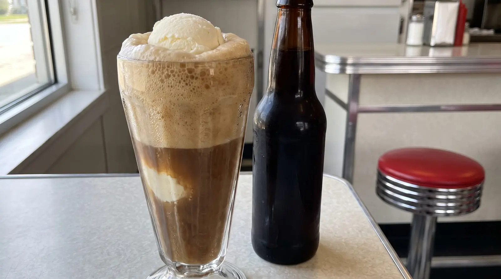 Root beer float in a tall vintage glass next to a dark glass bottle on a diner counter